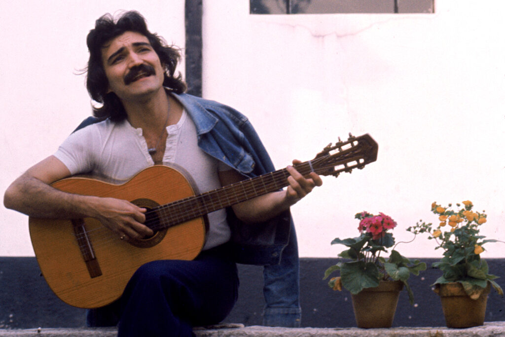 Foto do cantor Belchior tocando violão. Belchior é um homem de cabelos pretos e bigode cheio. Ele veste uma camiseta branca e tem um casaco jeans jogado sobre os ombros.