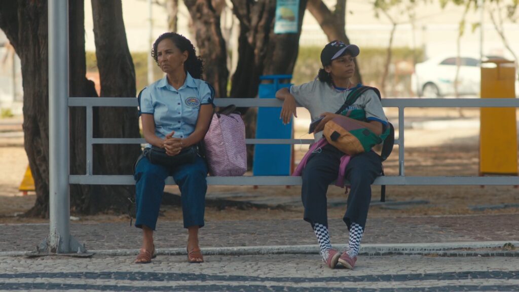 Cena do filme "Quando eu me encontrar". Na foto, mãe e filha, duas mulheres negras, estão sentadas em um ponto de ônibus, cada uma em uma ponta do banco, com um assento vazio entre elas.