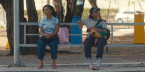 Cena do filme "Quando eu me encontrar". Na foto, mãe e filha, duas mulheres negras, estão sentadas em um ponto de ônibus, cada uma em uma ponta do banco, com um assento vazio entre elas.