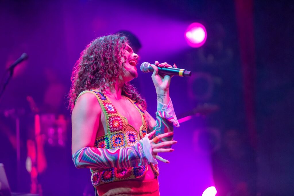 Imagem da cantora Makem, com roupa colorida e cabelos cacheados soltos, em cima do palco segurando um microfone enquanto canta. Ao fundo, estão alguns refletores que iluminam o palco com uma luz roxa.