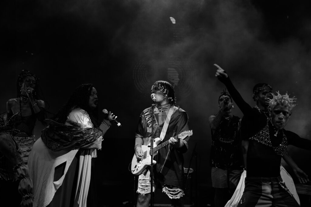 Imagem dos artistas Mateus Fazeno Rock e Jup do Bairro em preto e branco. Os dois estão olhando um para o outro enquanto cantam em seus microfones. Mateus também aparece tocando guitarra. Ao redor dos dois, dançarinos se apresentam.