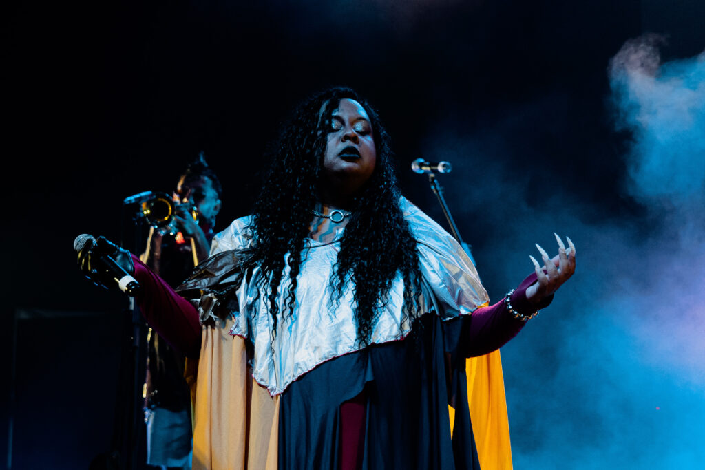 Imagem da cantora Jup do Bairro de braços abertos e olhos fechados. Ela está usando uma roupa com tons de prata, amarelo, azul e vermelho. Em uma de suas mãos, está segurando um microfone. Ao fundo, é possível ver uma fumaça azulada.